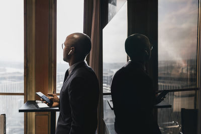 Side view of bald businessman wearing in-ear headphones holding smart phone looking away against television in board roo