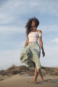 From below charming happy female in summer outfit walking along sandy hill on background of sundown looking away