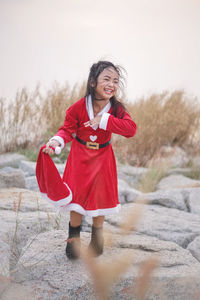 Cute girl wearing red dress holding santa hat while standing on rocks against sky