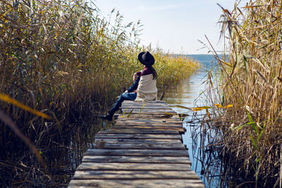 Woman in a black hat and sweater sits on the old pier to the bushes by the lake