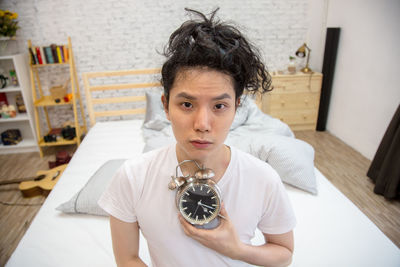 Portrait of young man holding alarm clock while sitting at home