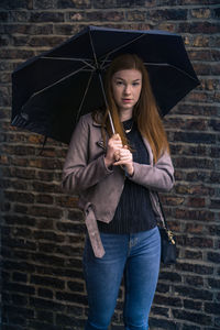 Portrait of young woman holding umbrella while standing against brick wall