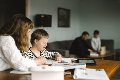 Boy with down syndrome reading book by mother while doing homework at table