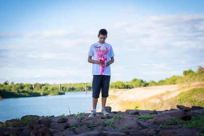 Young man holding bouquet while standing on rock against sky