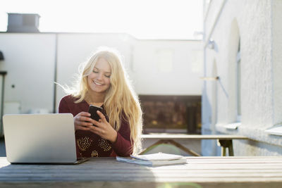 Happy teenage girl using mobile phone with laptop on table outdoors
