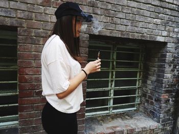 Side view of young woman smoking while standing by brick wall