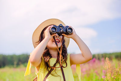 A happy cute girl stands in a field with high grass , looks into the sky through binoculars.