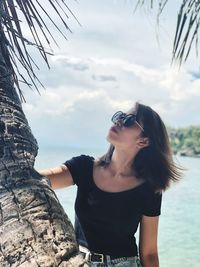 Beautiful young woman standing by tree trunk at beach against sky