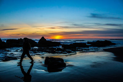 Side view of silhouette man with fishing rod walking on shore at beach against sky during sunset