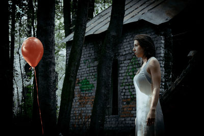 Woman looking at helium balloon while standing at forest