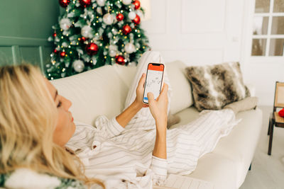 A woman lying on a couch in christmas uses a mobile phone for an investment application.