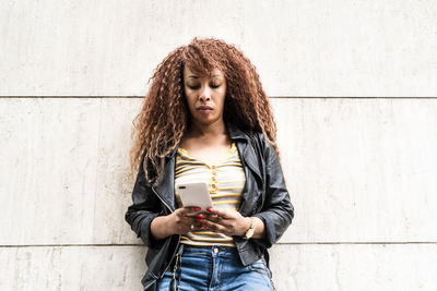 Young woman using phone while standing against wall