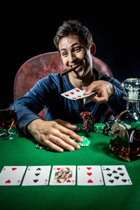 Smiling man sitting at poker table with whiskey and cigar against black background