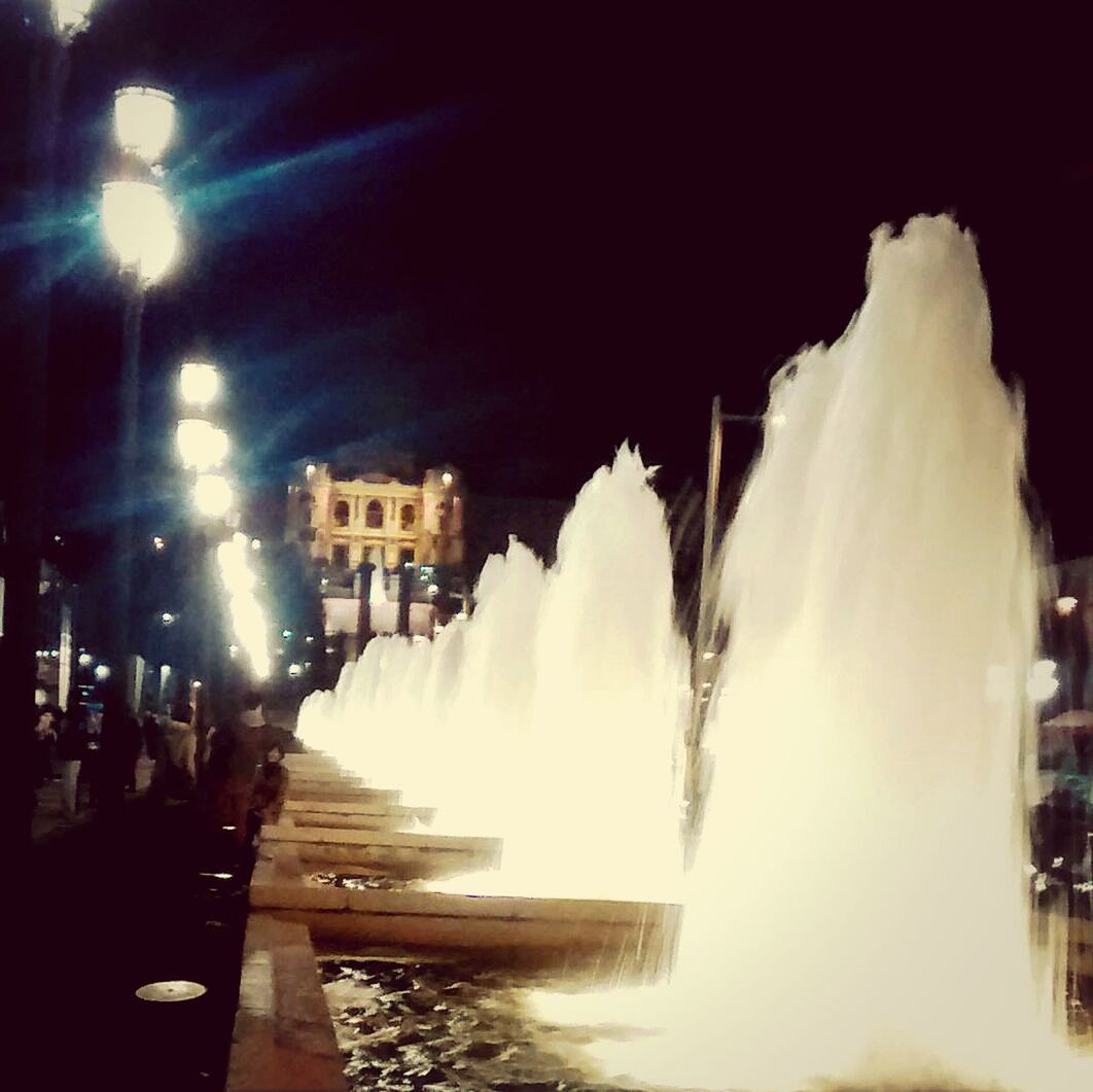 night, building exterior, illuminated, built structure, architecture, motion, city, long exposure, fountain, blurred motion, city life, sky, spraying, incidental people, outdoors, smoke - physical structure, winter, weather, cold temperature, street