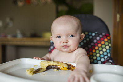 Portrait of shirtless baby girl with banana sitting on high chair