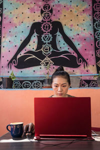 Serious asian female entrepreneur sitting at table and typing on netbook while working on project at home on background of wall with indian buddha poster