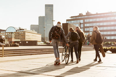 Rear view of friends with bicycle and skateboard on pedestrian zone in city against sky