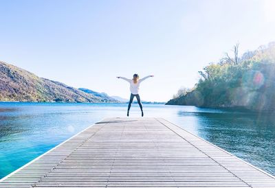 Rear view of woman jumping with arms outstretched on pier against lake