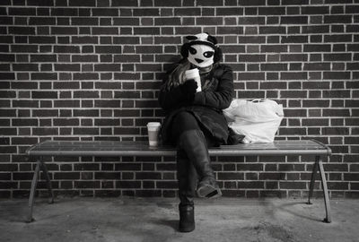 Young woman wearing mask while sitting on bench by brick wall