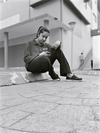 Young woman using phone while sitting on footpath in city