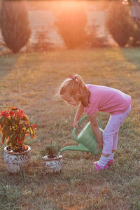 Girl watering plants while standing on grass against trees during sunset