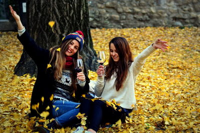 Smiling young friends having wine while sitting by trees during autumn