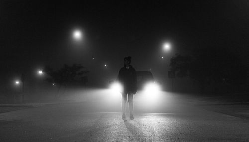 Woman standing against car with illuminated headlight on street at night