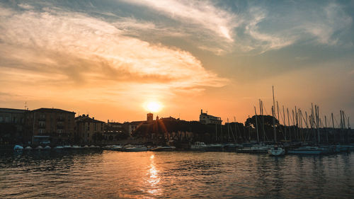 Sailboats moored in harbor by buildings against sky during sunset