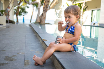 Little girl eating ice cream yogurt with spoon sitting by outdoor pool