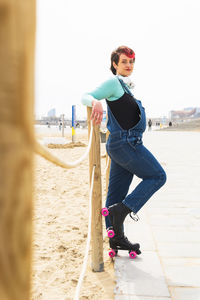 Full body of satisfied female in jeans overall and roller skates standing near wooden street fence and looking at camera