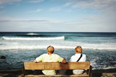 Rear view of women sitting on bench at beach against sky
