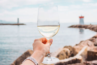 Woman hand with white wine glass on a sea, stones and lighthouse background. nice, french riviera
