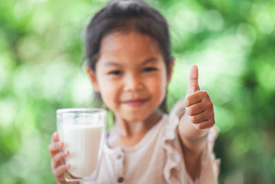 Portrait of girl gesturing thumbs up sign while holding milk glass outdoors