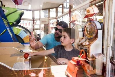 Father and son playing game at amusement arcade seen through window