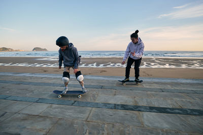 Young active male friends riding skateboards together along promenade on background of sea and sunset sky