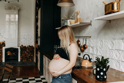 A young pregnant woman with a belly holding a mug drinking water standing in the kitchen at home