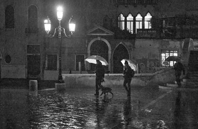 People with umbrella walking on wet illuminated street against buildings during rainy season