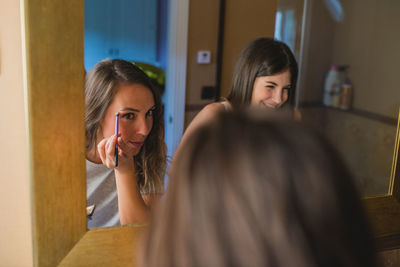 Delighted female friends standing in front of mirror at home and doing makeup together while preparing for party celebration