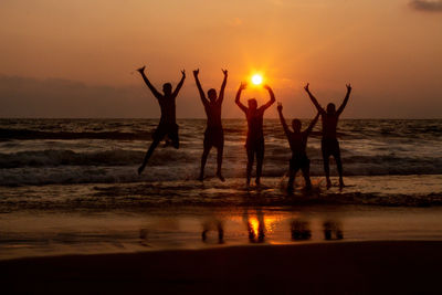 Silhouette friends with arms raised jumping at beach against sky during sunset