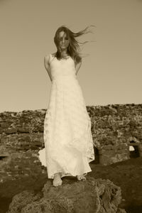 Sad young woman wearing white dress standing on rock against sky