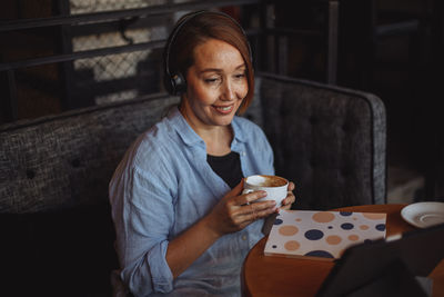 Middle-aged woman in a blue shirt works remotely on a laptop in a cafe
