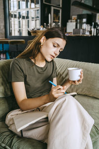 Young female entrepreneur writing in book holding coffee cup while sitting on sofa in studio