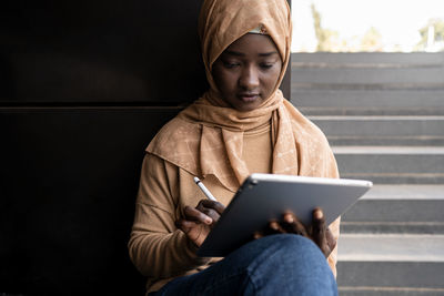 Concentrated young african american muslim female student in traditional headscarf sitting on street browsing on tablet with stylus while preparing for exams