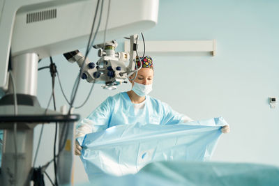 Attentive adult female medic in disposable mask and ornamental cap working in clinic with surgical microscope