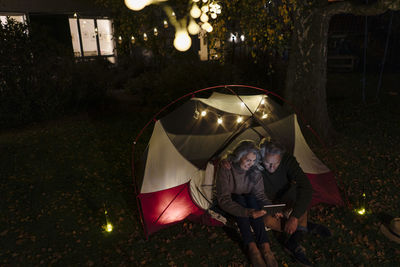 Senior couple with tablet camping in garden of their home at night