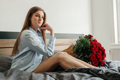 Beautiful girl is sitting on bed in bedroom with a huge bouquet of scarlet roses. flower delivery