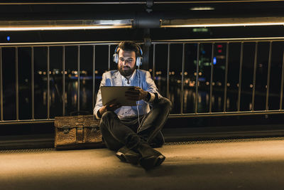 Smiling young man with tablet and headphones sitting on bridge at night