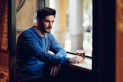 Thoughtful young man with drink looking through window in bar