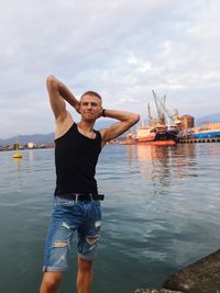 Young man with hands behind head standing on pier against sky