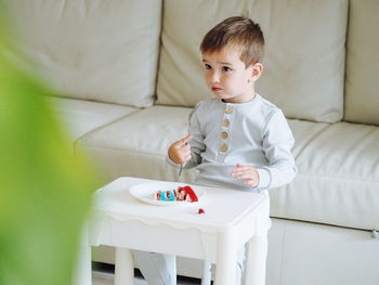 Cute toddler 3 years boy in grey pajamas eating red velvet cake in bright interior